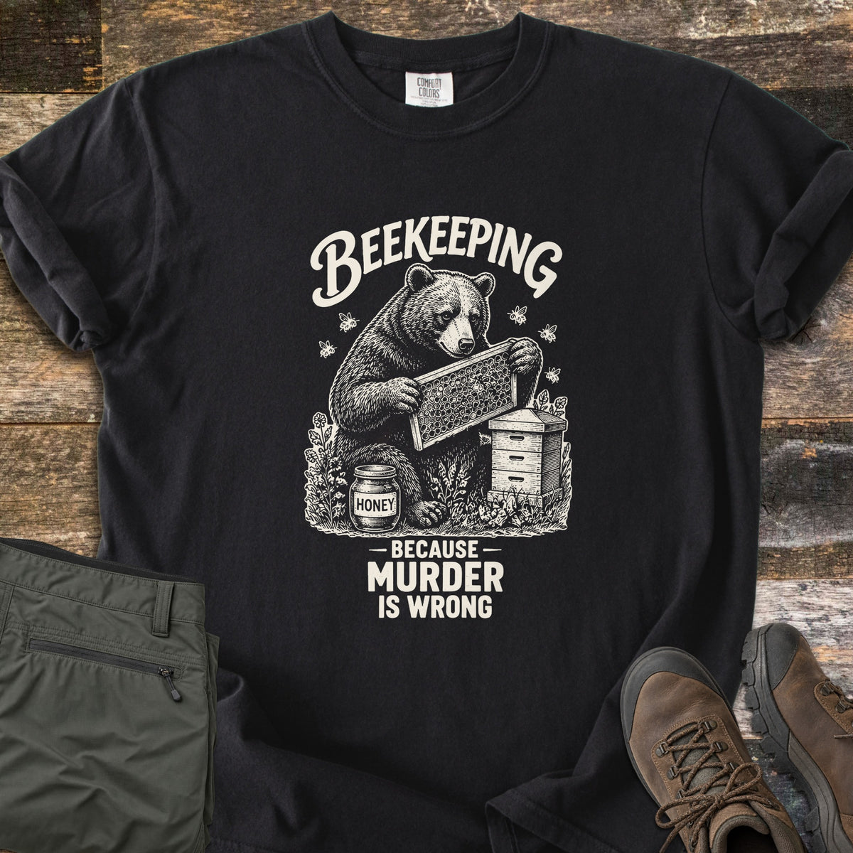 Beekeeping Because Murder Is Wrong Comfort Colors T-shirt in Black, laid flat. Cream ink artwork shows a bear holding a honeycomb frame beside a beehive and a jar labeled Honey, with bees around it, under the word Beekeeping and above Because Murder Is Wrong. Olive pants and brown hiking boots lie on wood.