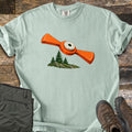 Winged Target Over the Pines Comfort Colors T-shirt in Bay, laid flat. An orange winged clay target with a white center flies over three pine trees on a mossy, rocky knoll, with no words. Folded olive hiking pants and brown hiking boots sit beside it on weathered wood planks.