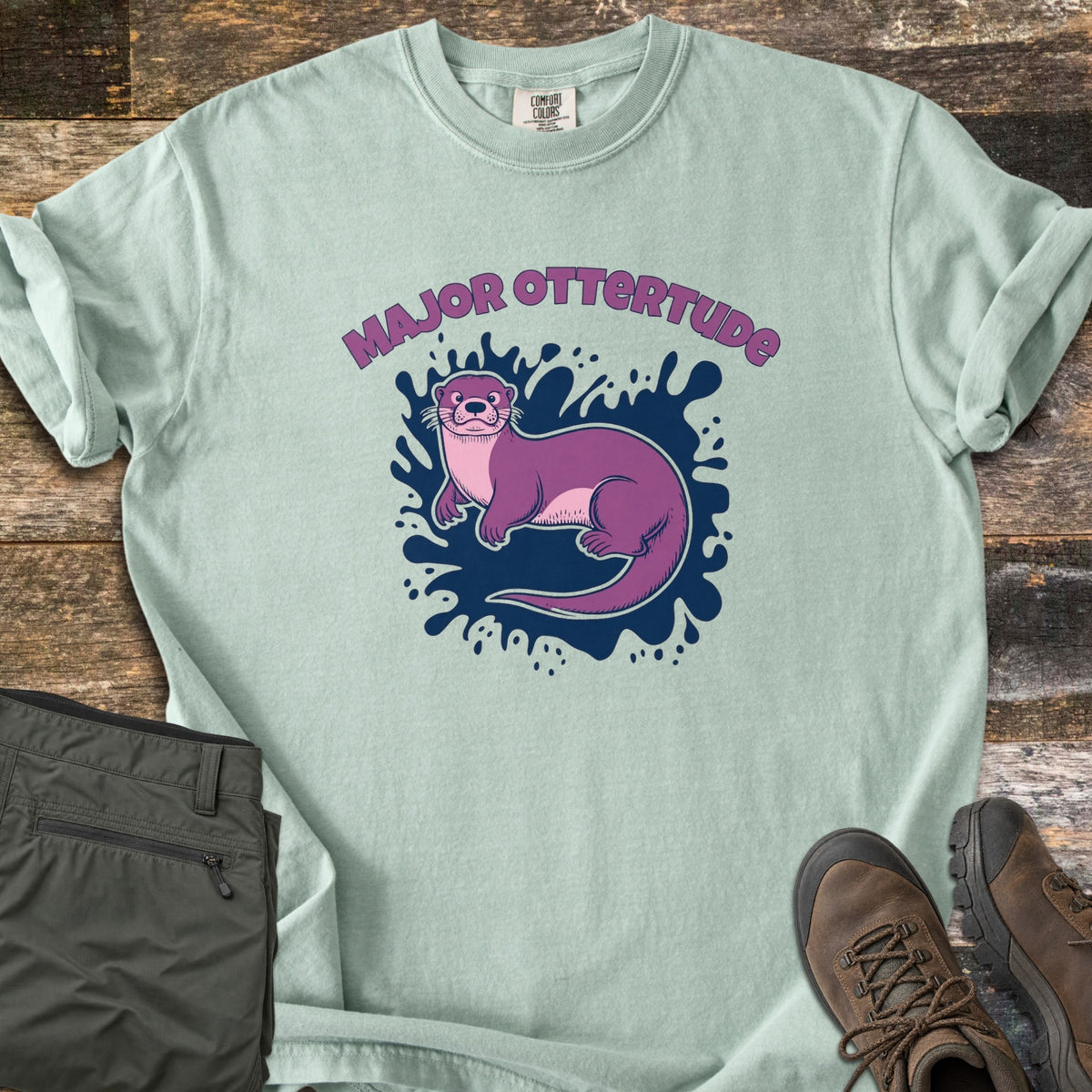 Major Ottertude Comfort Colors T-shirt in Bay, laid flat. A purple cartoon otter lies on a navy paint splash beneath the arched words Major Ottertude. Sleeves are rolled, on weathered wood with folded olive pants and brown hiking boots.