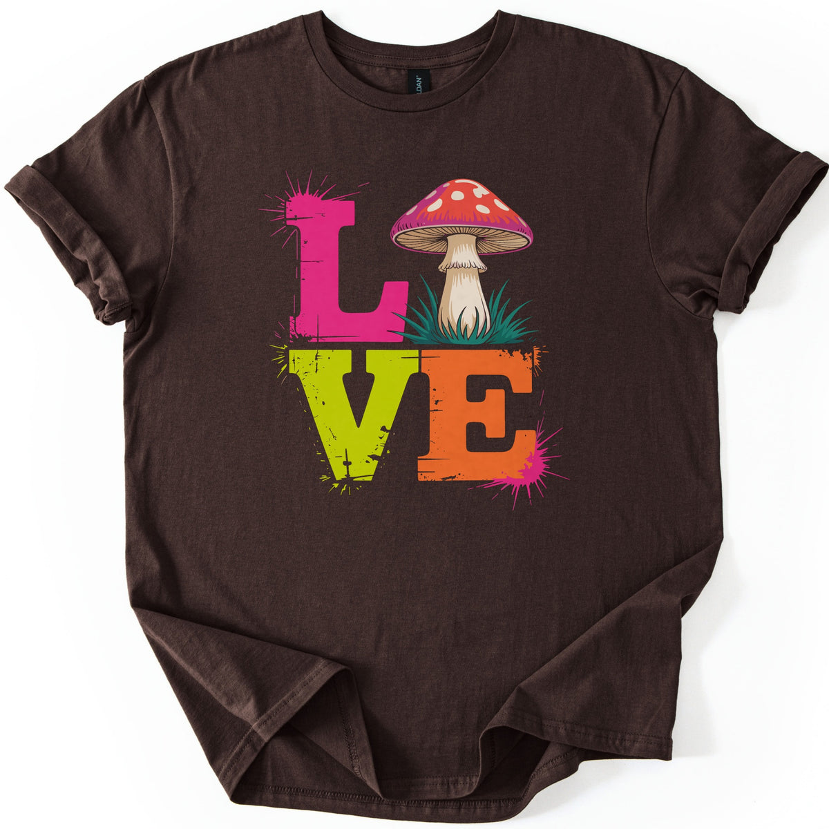 Mushroom Love Comfort Colors T-shirt in Dark Chocolate, laid flat. The word LOVE is stacked in distressed block letters: a pink L, a red mushroom with white spots in grass as the O, a lime green V and an orange E, with pink paint splatters. The sleeves are cuffed on a white background.