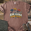 Ride Leader Herding Helmets Comfort Colors T-shirt in Espresso, laid flat. Three bike helmets in navy, yellow and orange linked by a winding trail to an arrow sign, above the words Ride Leader Herding Helmets MTB. Shown on weathered wood with olive pants and brown hiking boots.