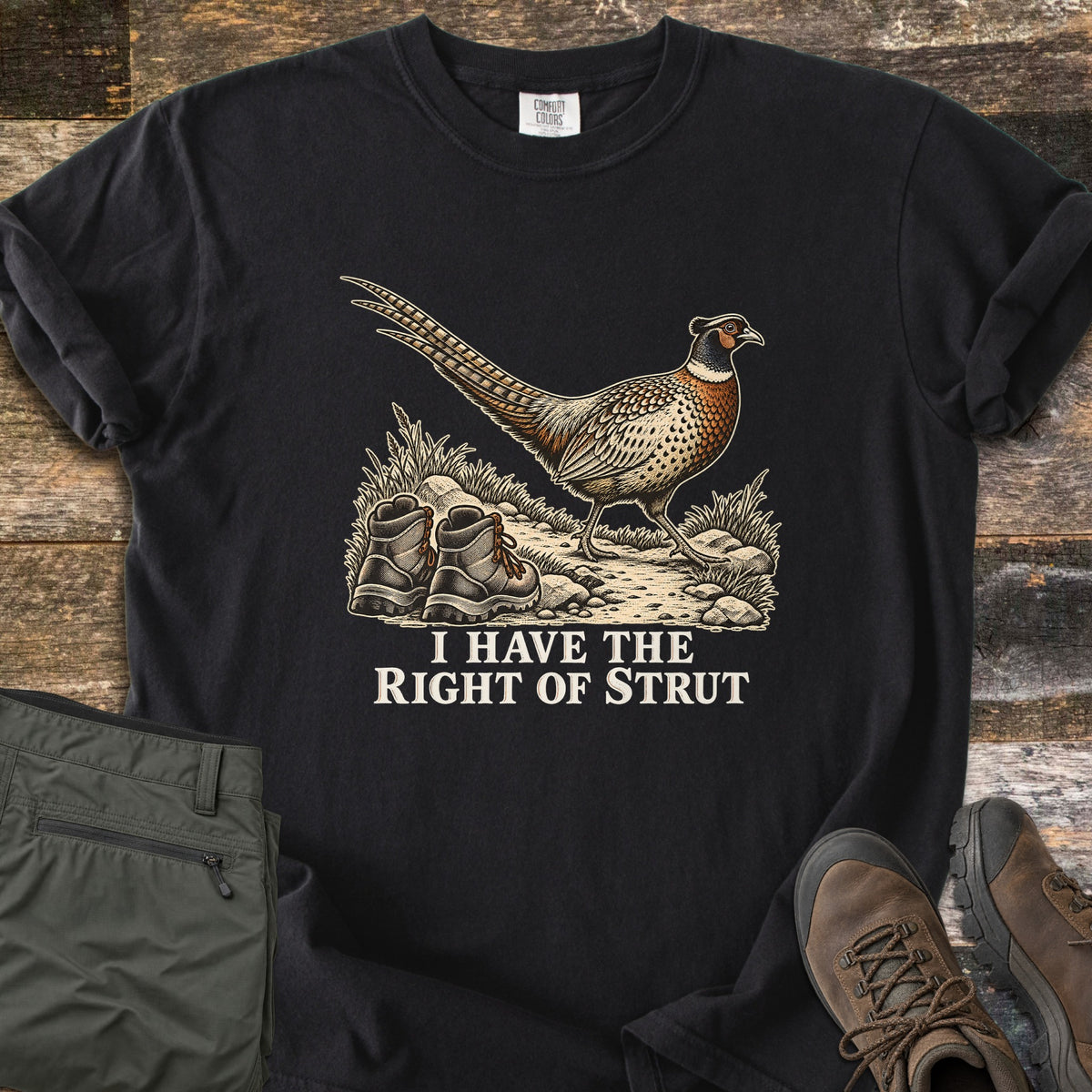 I Have the Right of Strut Comfort Colors T-shirt in Black, laid flat. A ring-necked pheasant walks a rocky trail past a pair of hiking boots above the words I Have the Right of Strut. Folded olive cargo pants and brown hiking boots lie beside it on weathered wood.