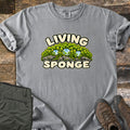 Living Sponge Comfort Colors T-shirt in Grey, laid flat. A cartoon mound of bright green moss on gray rock, holding three clear water droplets, between the words Living Sponge. Olive hiking pants and brown leather hiking boots lie beside it on weathered wood.