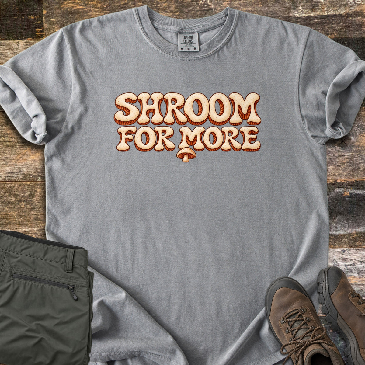 Shroom for More Comfort Colors T-shirt in Grey, laid flat. Retro cream bubble letters with rust-orange striped shadows read Shroom For More, with a small mushroom below. Sleeves are rolled on a rustic wood floor beside olive cargo pants and brown hiking boots.