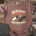 Sealant Confetti Unplanned Party Comfort Colors T-shirt in Espresso, laid flat. A cream sealant bottle squirts toward a knobby bike tire amid orange and green dots, with the words Sealant Confetti Unplanned Party MTB. Folded olive pants and brown hiking boots sit on weathered wood.