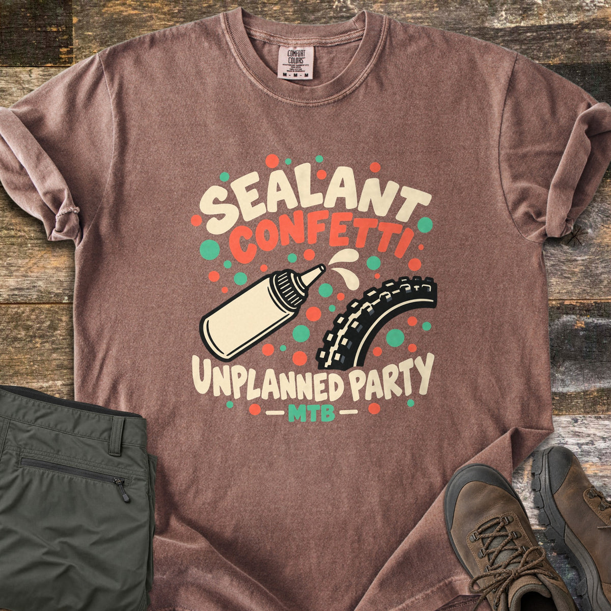 Sealant Confetti Unplanned Party Comfort Colors T-shirt in Espresso, laid flat. A cream sealant bottle squirts toward a knobby bike tire amid orange and green dots, with the words Sealant Confetti Unplanned Party MTB. Folded olive pants and brown hiking boots sit on weathered wood.