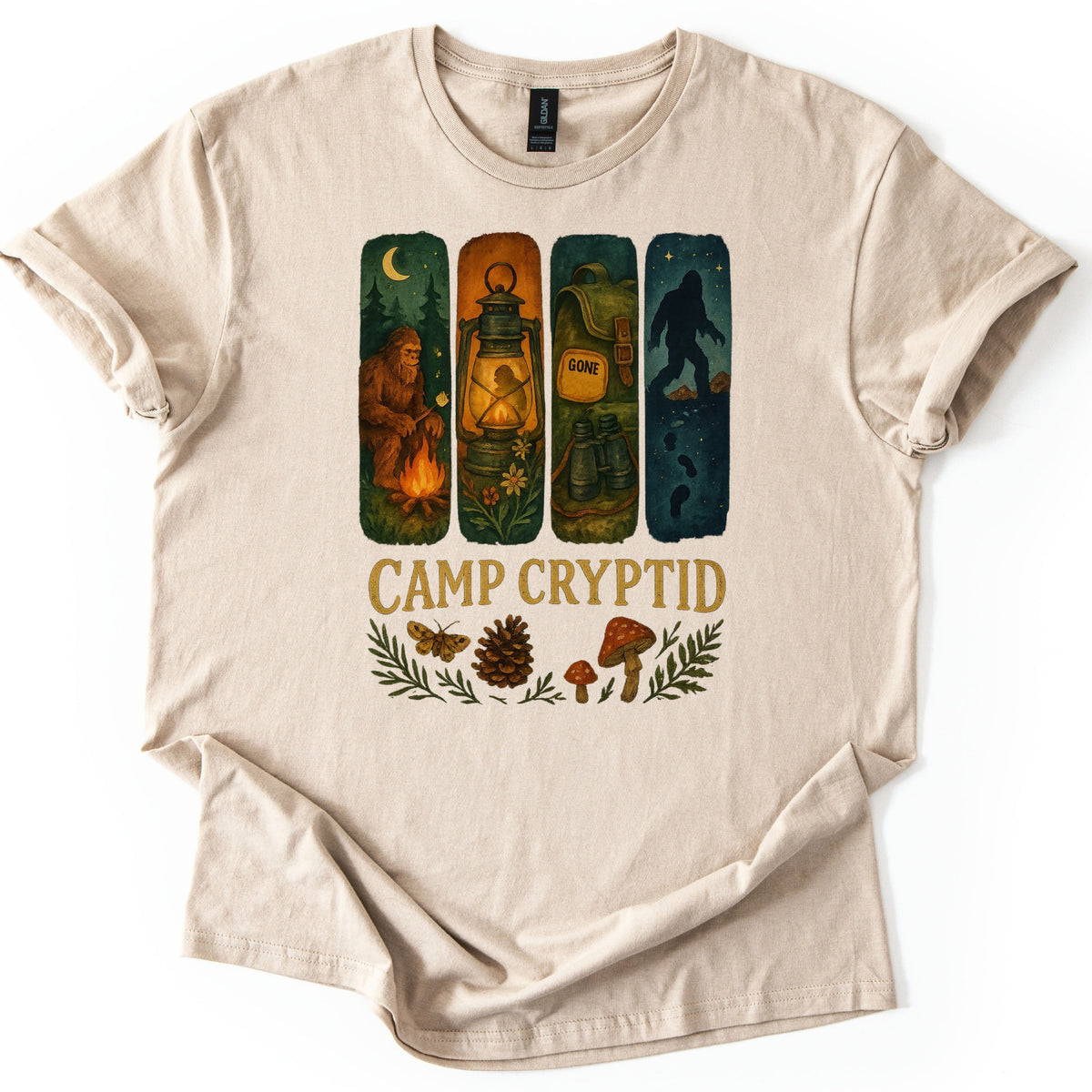 Camp Cryptid Big Foot Comfort Colors T-shirt in Sand, laid flat. Four painted panels show Bigfoot at a campfire, a lit lantern, a backpack marked GONE, and Bigfoot's silhouette under stars, above the words Camp Cryptid and a moth, pinecone and mushrooms. Shown on a white background.