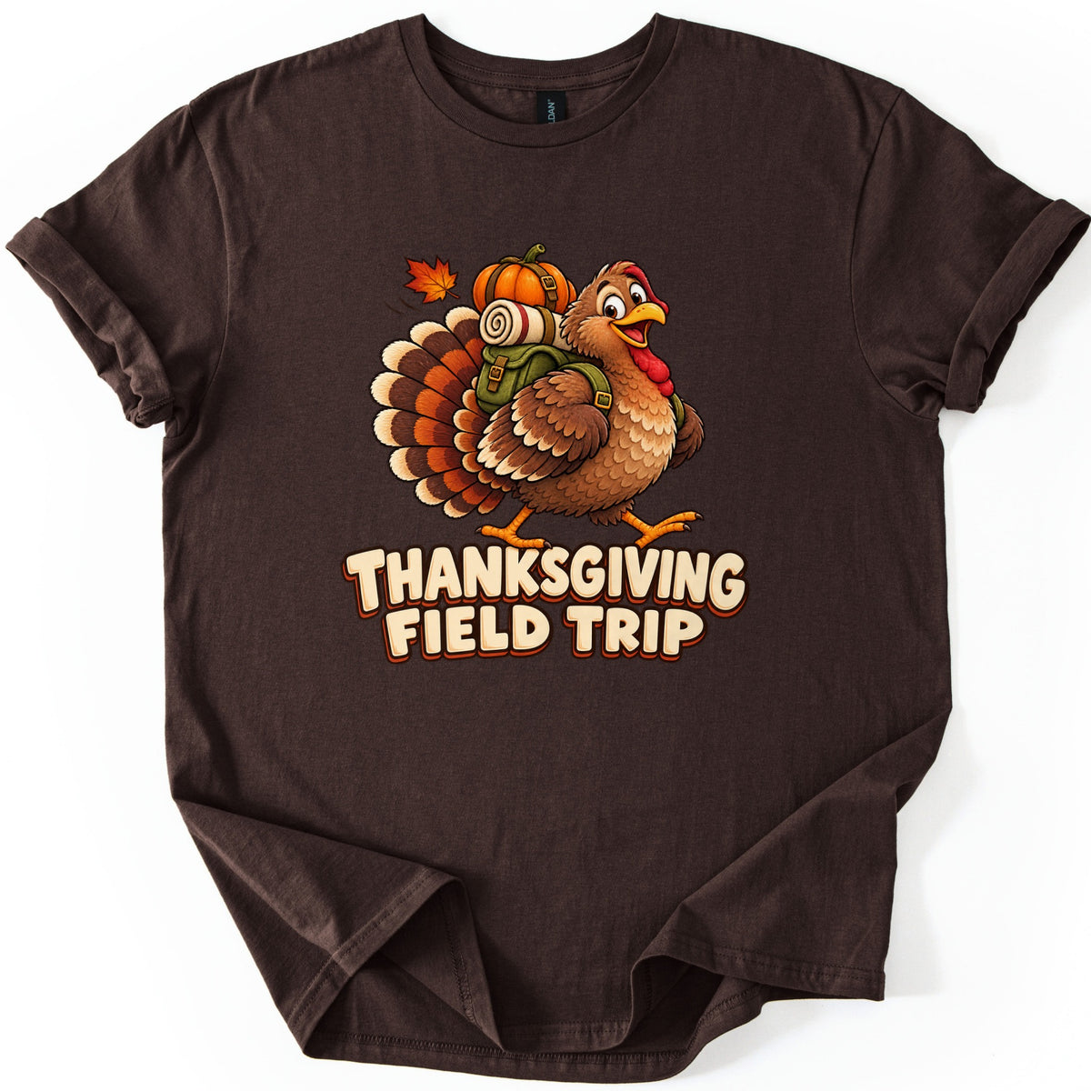 Thanksgiving Field Trip Comfort Colors T-shirt in Dark Chocolate, laid flat. A smiling cartoon turkey walks with a green backpack topped by a rolled blanket and a pumpkin, beside a falling maple leaf, above the words Thanksgiving Field Trip. The sleeves are cuffed on a white surface.