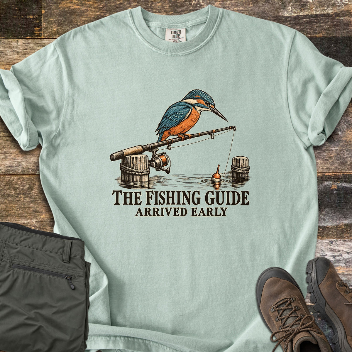 The Fishing Guide Arrived Early Comfort Colors T-shirt in Bay, laid flat. A kingfisher perches on a fishing rod resting on rope-wrapped dock posts, its line and orange bobber in the water, above the words The Fishing Guide Arrived Early. Olive pants and brown hiking boots lie on weathered wood.