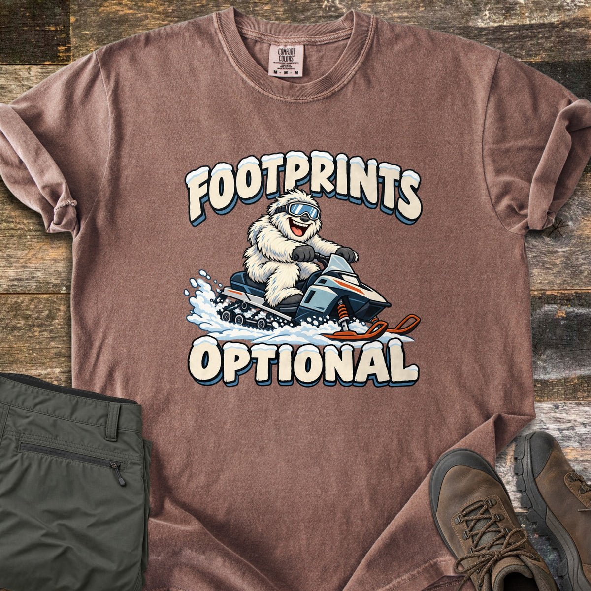 Footprints Optional Comfort Colors T-shirt in Espresso, laid flat. A grinning white yeti in ski goggles rides a snowmobile through spraying snow between the snow-capped words Footprints Optional. It lies on weathered wood beside folded olive hiking pants and brown hiking boots.