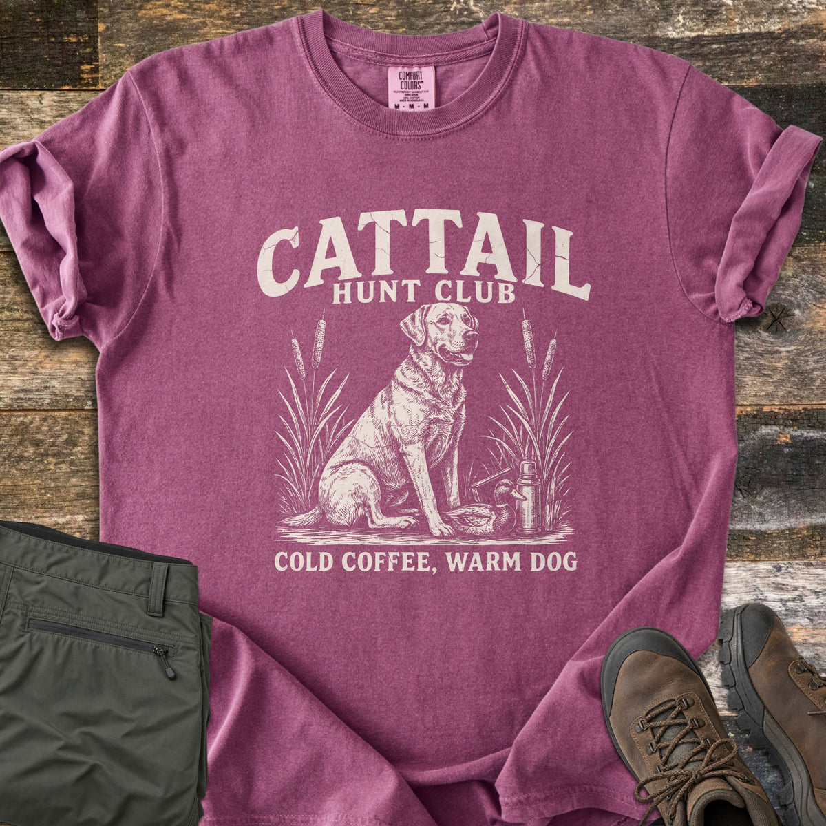 Cattail Hunt Club Comfort Colors T-shirt in Berry, laid flat. Cream print of a Labrador sitting among cattails beside a duck decoy and thermos, under the words Cattail Hunt Club, above Cold Coffee, Warm Dog. On weathered wood with olive pants and brown hiking boots.