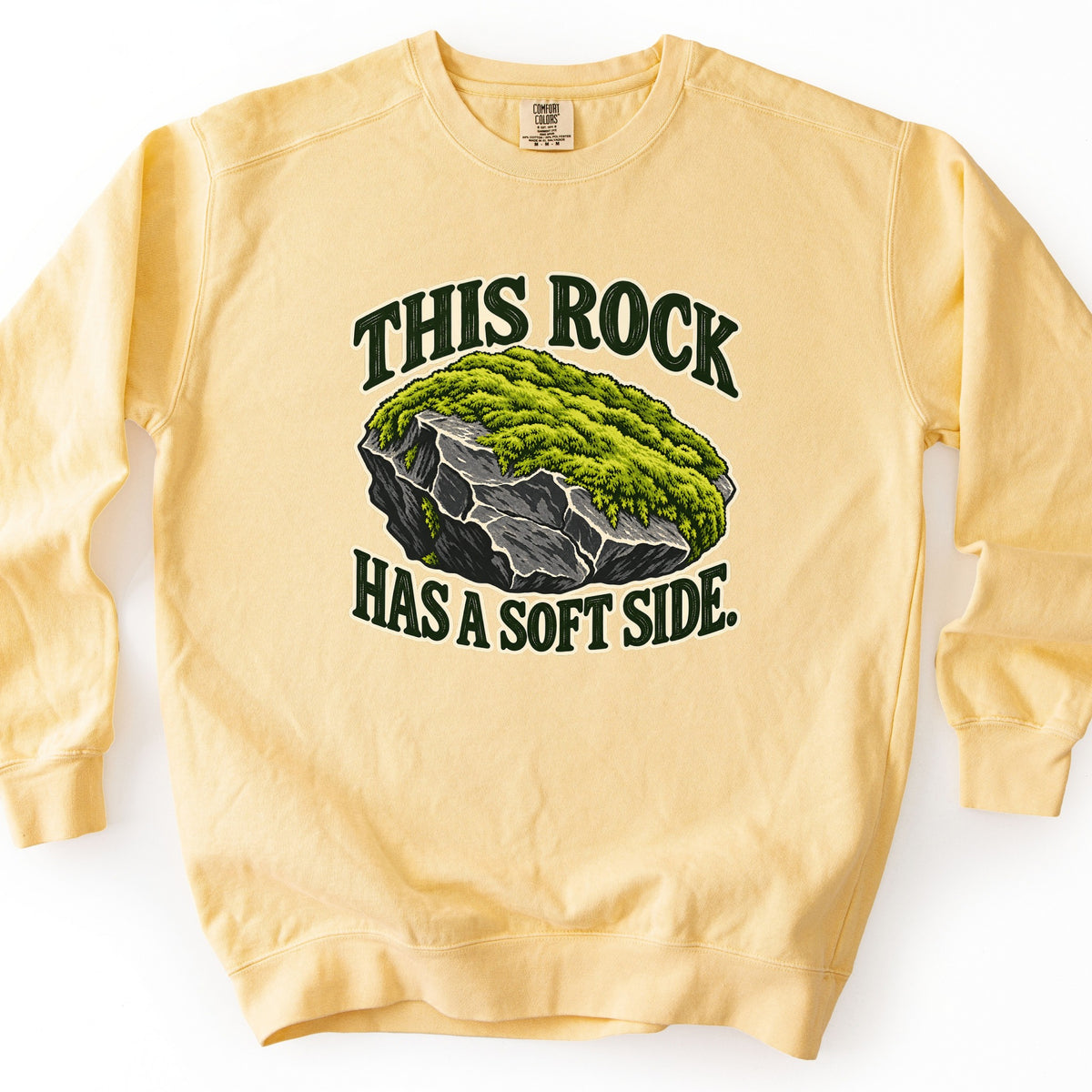 This Rock Has a Soft Side Comfort Colors crewneck sweatshirt in Butter, laid flat. A gray boulder topped with bright green moss sits between the words This Rock above and Has a Soft Side. below in dark green letters. The sweatshirt lies on a plain white surface, sleeves out.