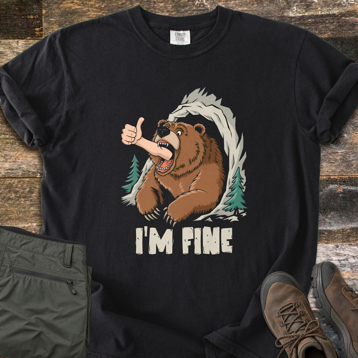I'm Fine Comfort Colors T-shirt in Black, laid flat. A cartoon brown bear bursts from a rocky cave framed by pine trees, a human arm in its mouth giving a thumbs-up, above the words I'M FINE. Olive pants and brown hiking boots sit on weathered wood.