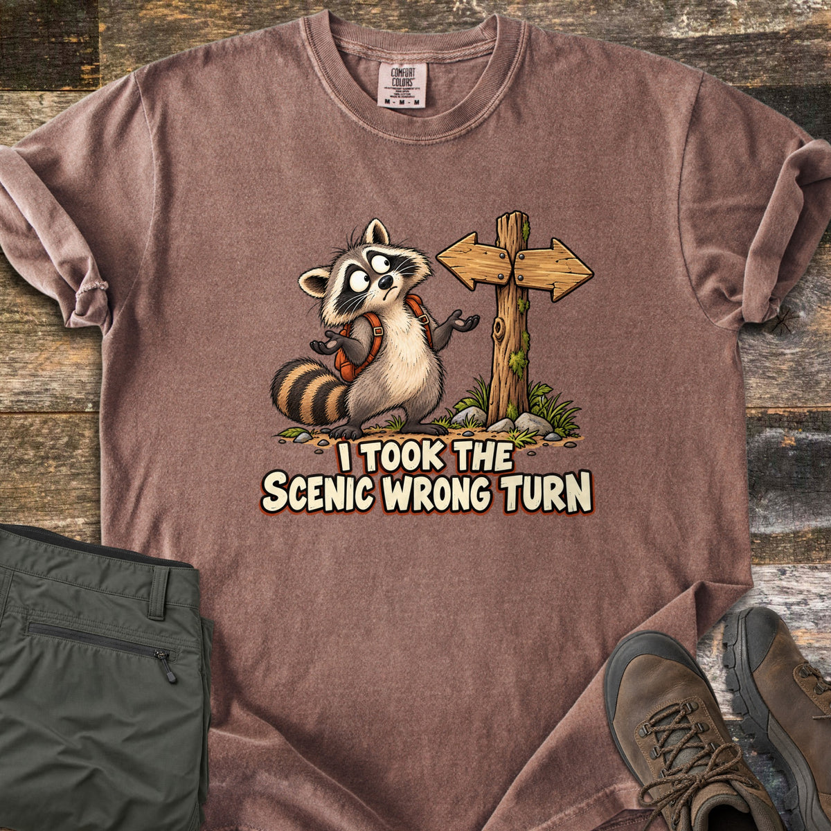 I Took the Scenic Wrong Turn Comfort Colors T-shirt in Espresso, laid flat. A cartoon raccoon with a backpack shrugs, looking confused beside a wooden signpost with arrows pointing both ways, above the words I Took the Scenic Wrong Turn. Olive hiking pants and brown boots lie on weathered wood.