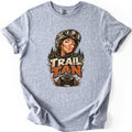 Trail Tan Comfort Colors T-shirt in Sport Grey, laid flat. Front print shows a smiling woman with a mud-splattered face, helmet and goggles, pine trees behind her, above the words TRAIL TAN and a muddy ATV. The shirt lies on a white background.
