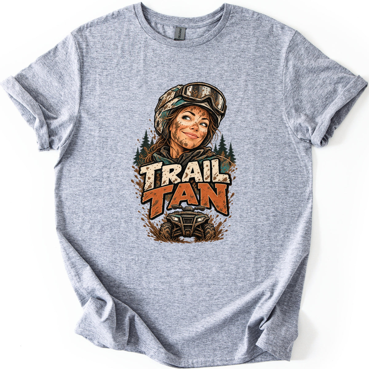 Trail Tan Comfort Colors T-shirt in Sport Grey, laid flat. Front print shows a smiling woman with a mud-splattered face, helmet and goggles, pine trees behind her, above the words TRAIL TAN and a muddy ATV. The shirt lies on a white background.