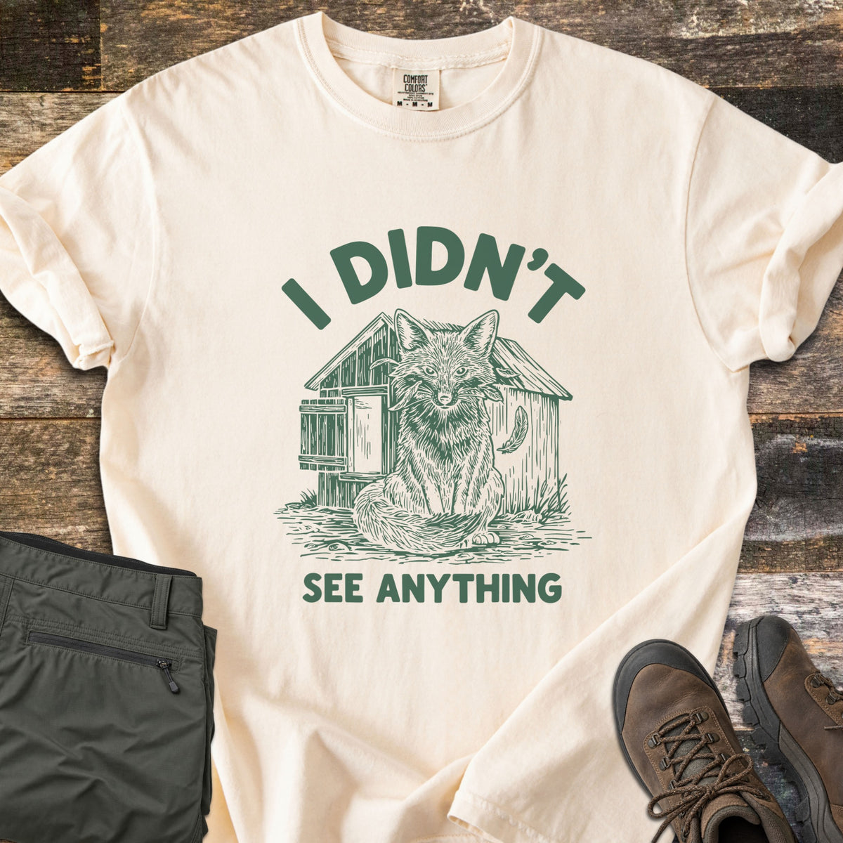 I Didn't See Anything Comfort Colors T-shirt in Ivory, laid flat. A green sketch of a fox with a feather in its mouth sits before a wooden chicken coop, between the words I Didn't and See Anything. Folded olive pants and brown hiking boots lie beside it on dark wood.