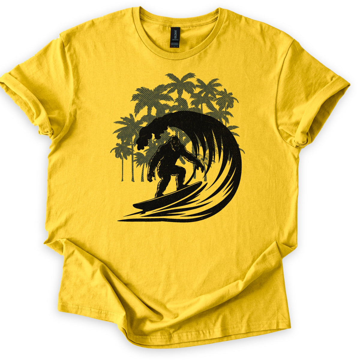 Surf Squatch Comfort Colors T-shirt in Daisy, laid flat. A black silhouette of Bigfoot riding a surfboard through a curling wave, with olive green halftone palm trees behind him. No words are printed. The shirt has rolled sleeves on a white background.