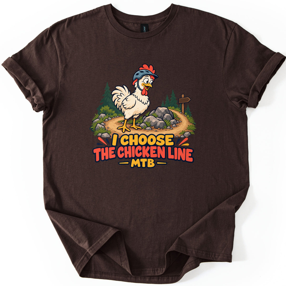 I Choose the Chicken Line MTB Comfort Colors T-shirt in Dark Chocolate, laid flat. A cartoon chicken in a bike helmet stands on a rocky dirt trail with pine trees and a wooden sign above the words I Choose the Chicken Line MTB. The shirt has rolled sleeves on a white background.