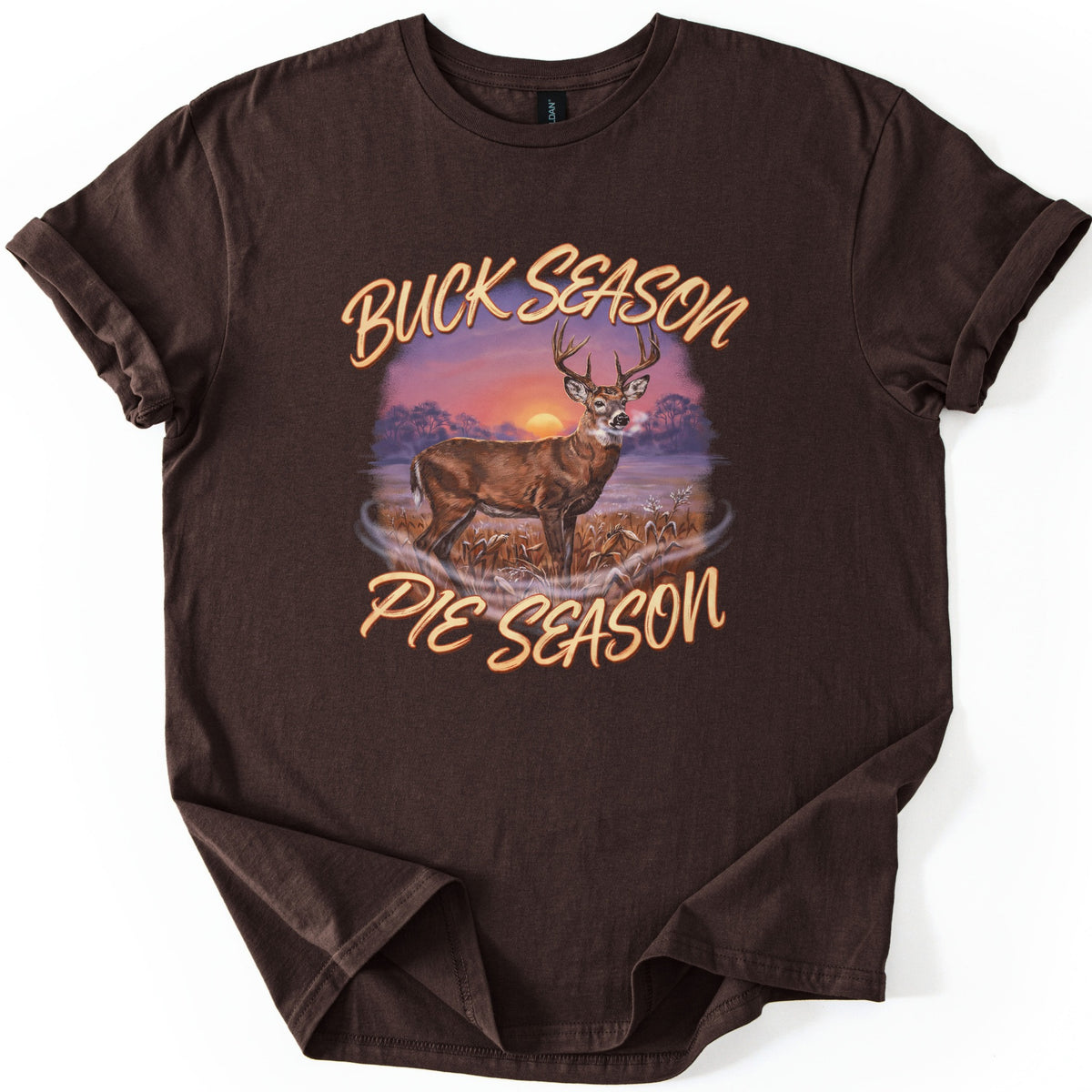 Buck Season Pie Season Comfort Colors T-shirt in Dark Chocolate, laid flat. A painted whitetail buck stands in a frosty cornfield under a purple sunset, framed by the words Buck Season and Pie Season in peach script. The sleeves are cuffed, on a white background.