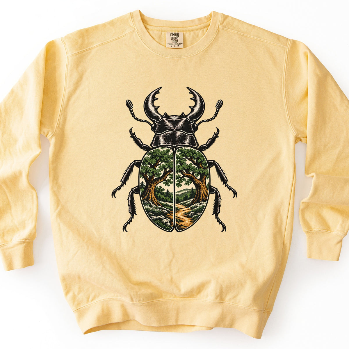 Beetle Forest Path Comfort Colors crewneck sweatshirt in Butter, laid flat. A black stag beetle with large antler-like jaws is printed on the front. Its wing covers show two oak trees arching over a winding dirt path toward green hills. No words are printed. It lies on white.