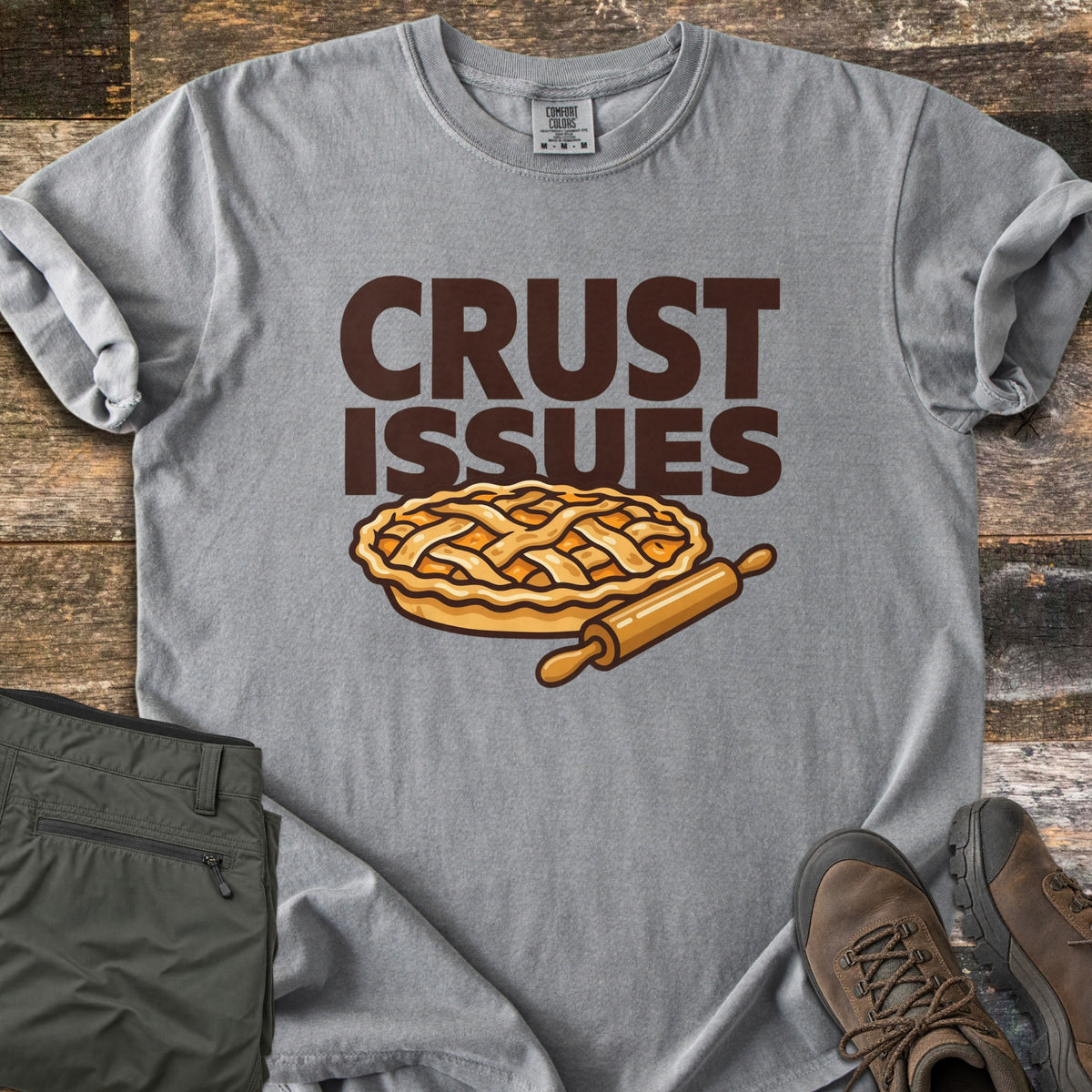 Crust Issues Comfort Colors T-shirt in Grey, laid flat. The words CRUST ISSUES in dark brown block letters sit above a cartoon lattice-top pie and a wooden rolling pin. Olive pants and brown hiking boots lie beside it on a rustic wood surface.