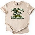 Look Closer Tread Lighter Comfort Colors T-shirt in Sand, laid flat. A dark green hiking boot with gold laces hovers above a mound of green moss, between the words Look Closer and Tread Lighter in curvy dark green letters. The shirt lies on a white background with cuffed sleeves.