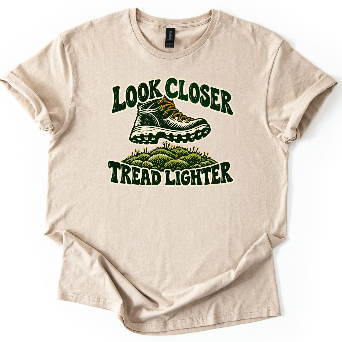 Look Closer Tread Lighter Comfort Colors T-shirt in Sand, laid flat. A dark green hiking boot with gold laces hovers above a mound of green moss, between the words Look Closer and Tread Lighter in curvy dark green letters. The shirt lies on a white background with cuffed sleeves.
