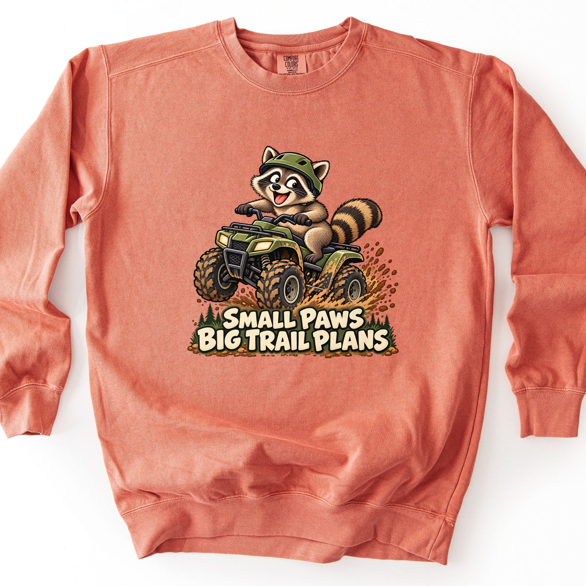 Small Paws Big Trail Plans Comfort Colors crewneck sweatshirt in Terracotta, laid flat. A grinning cartoon raccoon in a green helmet rides a muddy green ATV, kicking up mud past pine trees, above the words Small Paws Big Trail Plans. The sweatshirt lies on a white background.