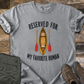 Reserved For My Favorite Human Comfort Colors T-shirt in Grey, laid flat. A yellow canoe seen from above with a red heart on each seat sits between two red-bladed paddles, under the words Reserved For and above My Favorite Human. Olive pants and brown hiking boots lie beside it on wood planks.