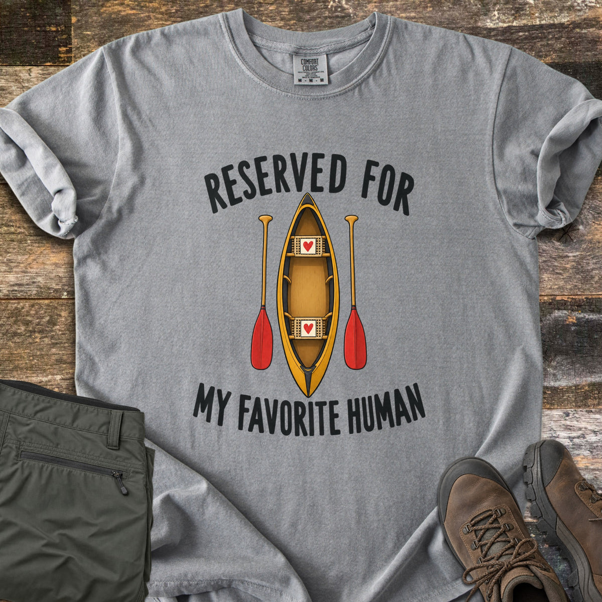 Reserved For My Favorite Human Comfort Colors T-shirt in Grey, laid flat. A yellow canoe seen from above with a red heart on each seat sits between two red-bladed paddles, under the words Reserved For and above My Favorite Human. Olive pants and brown hiking boots lie beside it on wood planks.