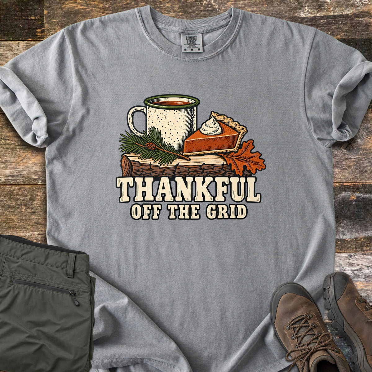 Thankful Off the Grid Comfort Colors T-shirt in Grey, laid flat. A speckled enamel camp mug and a slice of pumpkin pie with whipped cream sit on a log with a pine sprig and an oak leaf above the words Thankful Off the Grid. Olive hiking pants and brown boots lie on weathered wood.