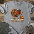 Migration Plans Changed for November Comfort Colors T-shirt in Grey, laid flat. A cartoon turkey looks through binoculars beside a wooden arrow sign and a small pumpkin above the words Migration Plans Changed for November. Shown on rustic wood with olive pants and brown hiking boots.