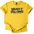 Walked It Still Counts Comfort Colors T-shirt in Daisy, laid flat. Navy brush-stroke letters read Walked It Still Counts, with a rust-orange dashed trail line underneath ending in the small letters MTB. The shirt has rolled sleeves and lies on a plain white background.