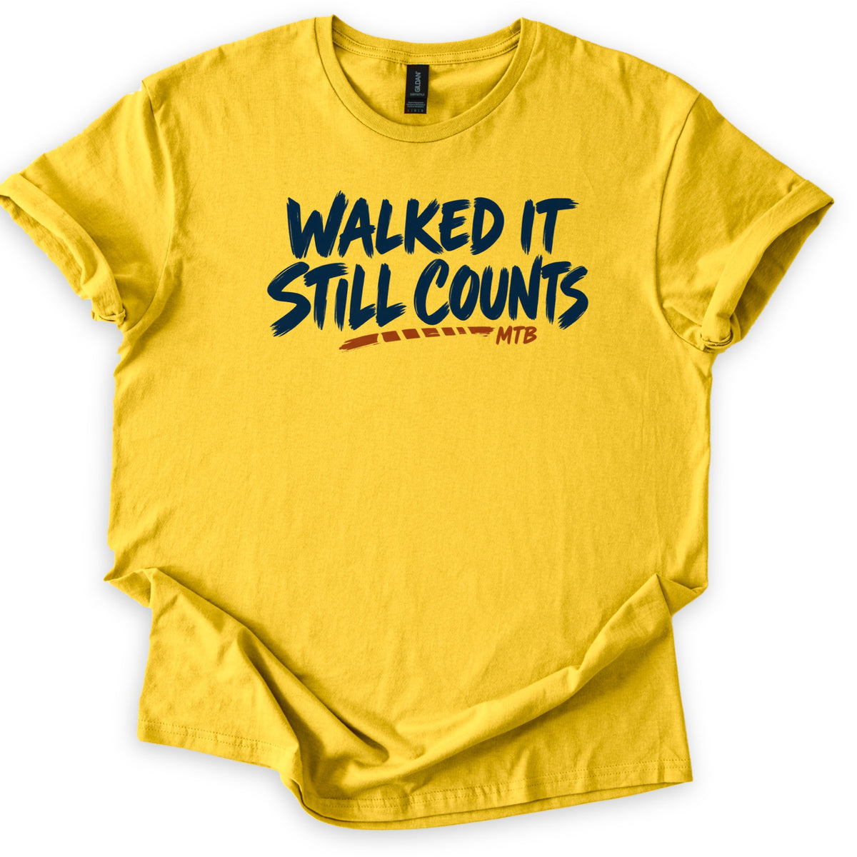 Walked It Still Counts Comfort Colors T-shirt in Daisy, laid flat. Navy brush-stroke letters read Walked It Still Counts, with a rust-orange dashed trail line underneath ending in the small letters MTB. The shirt has rolled sleeves and lies on a plain white background.