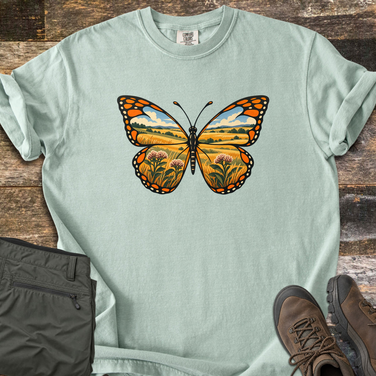 Monarch Prairie Comfort Colors T-shirt in Bay, laid flat. A monarch butterfly whose wings frame a prairie scene of golden grass, pink milkweed blooms and a blue sky with clouds. Olive cargo pants and brown hiking boots sit beside it on weathered wood planks.