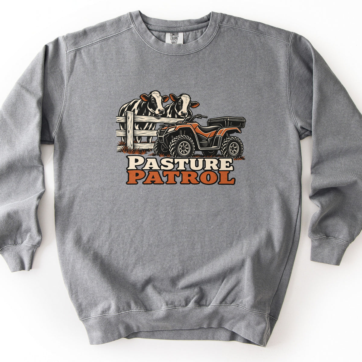 Pasture Patrol Comfort Colors crewneck sweatshirt in Grey, laid flat. Two black-and-white cows peer over a wooden fence beside an orange and black four-wheeler, above the words Pasture Patrol in cream and rust orange. The sweatshirt lies on a plain white surface.