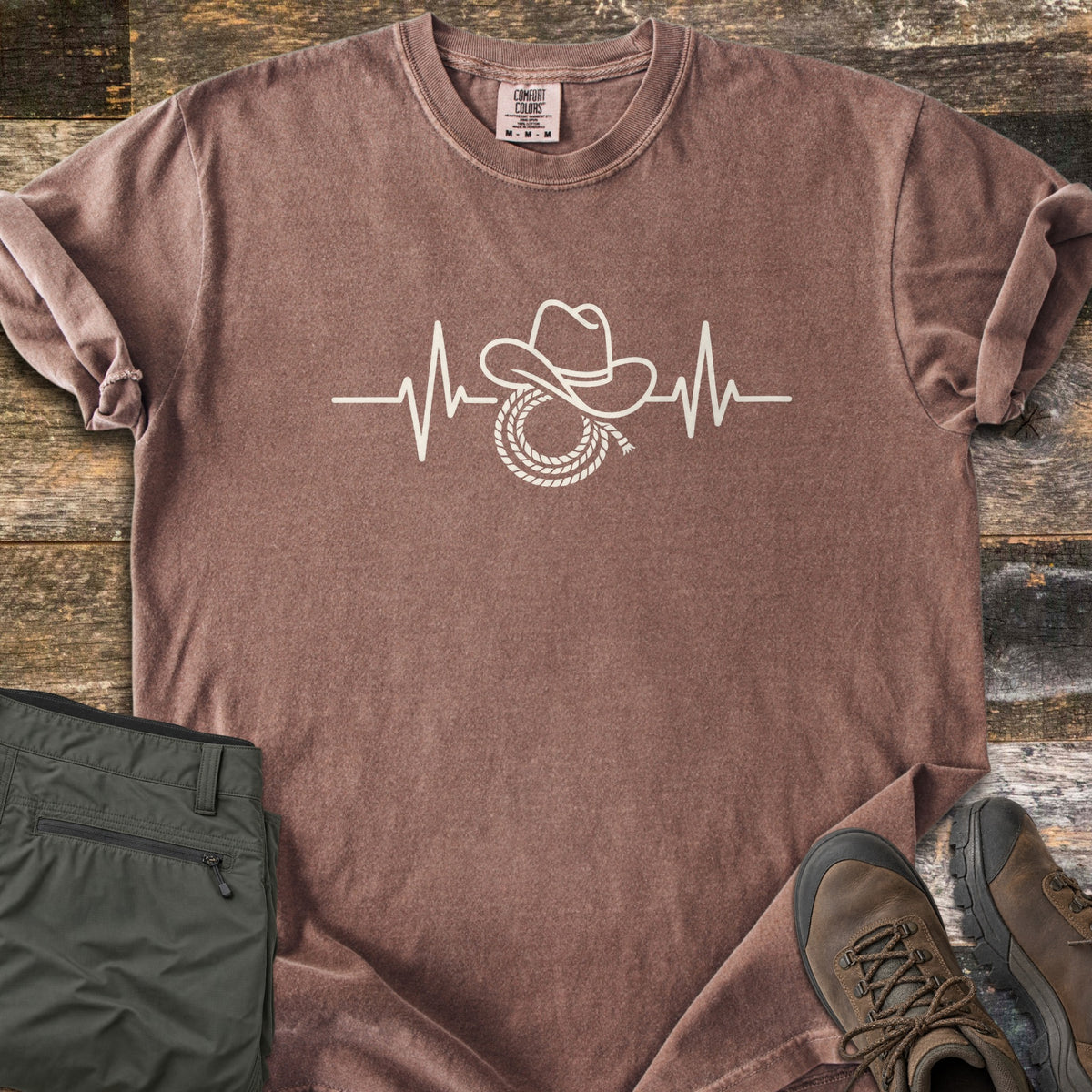 Cowboy Heartbeat Comfort Colors T-shirt in Espresso, laid flat. A cream line art heartbeat line rises into peaks around a cowboy hat resting on a coiled lasso; there are no words. Sleeves are cuffed, on rustic wood boards beside folded olive cargo pants and brown hiking boots.