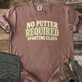 No Putter Required Comfort Colors T-shirt in Espresso, laid flat. Wavy block letters read No Putter Required in cream and sage green, with Sporting Clays below. Sleeves are rolled on a rustic wood surface beside folded olive cargo pants and brown leather hiking boots.