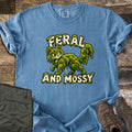 Feral and Mossy Comfort Colors T-shirt in Blue Jean, laid flat. A cartoon cat made of green moss with bits of bark walks between the words Feral and Mossy in cream letters dripping with moss. Folded olive pants and brown hiking boots sit on a weathered wood floor.
