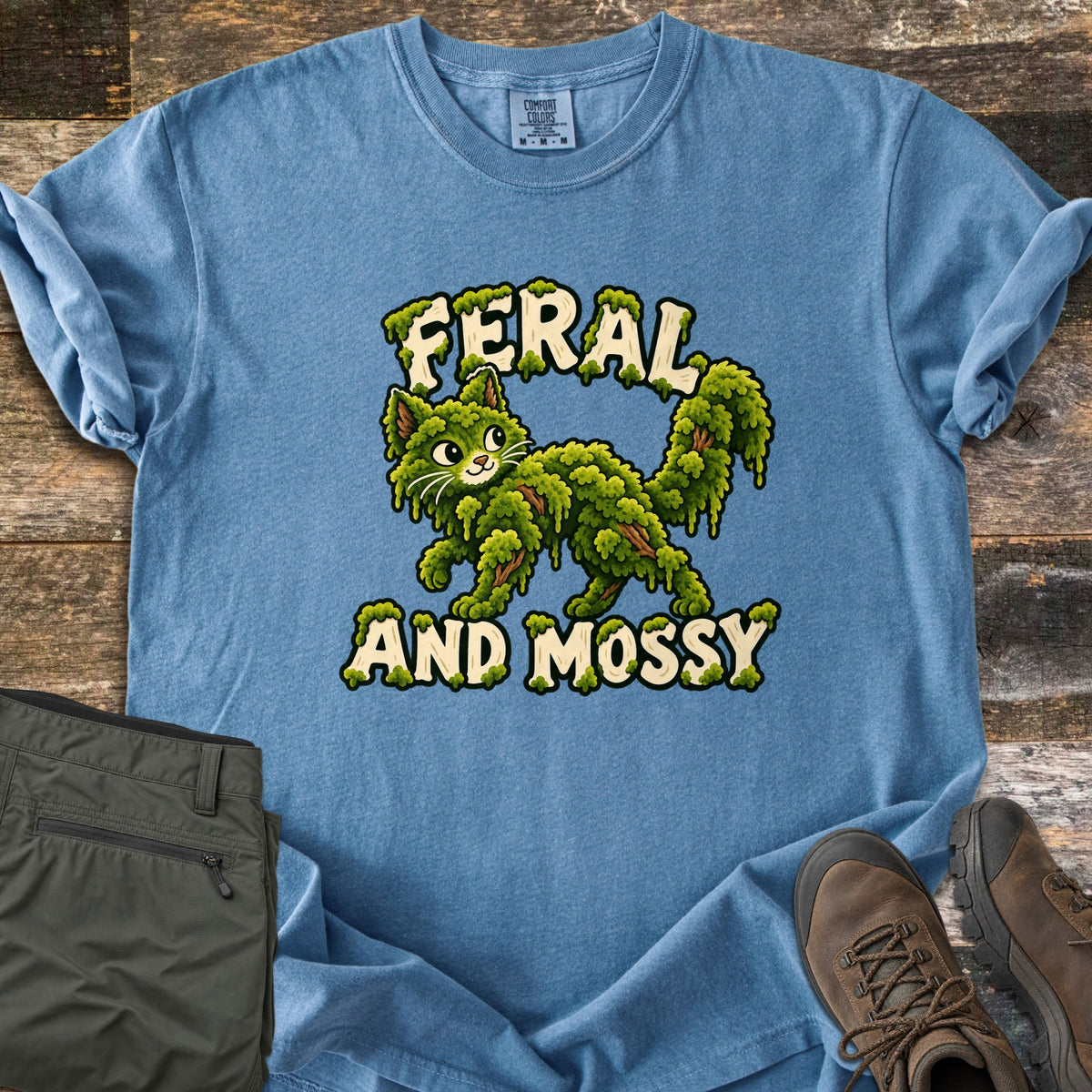 Feral and Mossy Comfort Colors T-shirt in Blue Jean, laid flat. A cartoon cat made of green moss with bits of bark walks between the words Feral and Mossy in cream letters dripping with moss. Folded olive pants and brown hiking boots sit on a weathered wood floor.