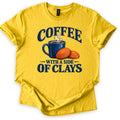 Coffee with a Side of Clays Comfort Colors T-shirt in Daisy, laid flat. A steaming navy enamel mug of coffee sits beside two orange clay targets, with the words COFFEE above and WITH A SIDE OF CLAYS below in navy. The shirt lies on a white background with cuffed sleeves.