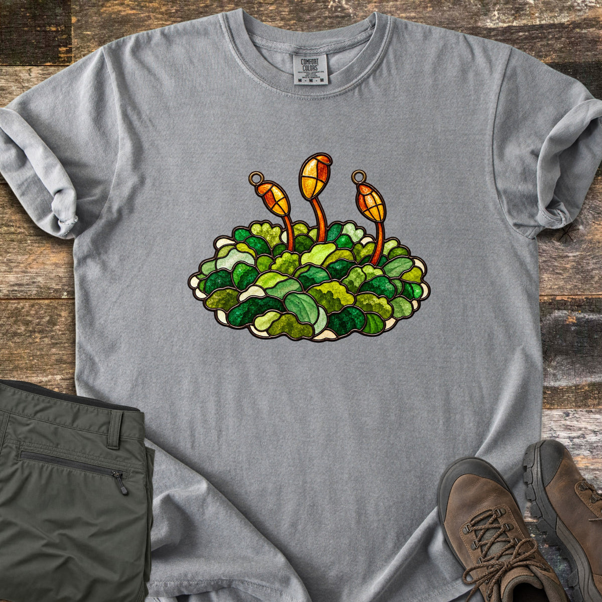 Stained Glass Moss Comfort Colors T-shirt in Grey, laid flat. A stained glass style mound of green moss with three orange and gold spore capsules on red stalks, with no words. Folded olive pants and brown hiking boots sit beside it on weathered wood planks.