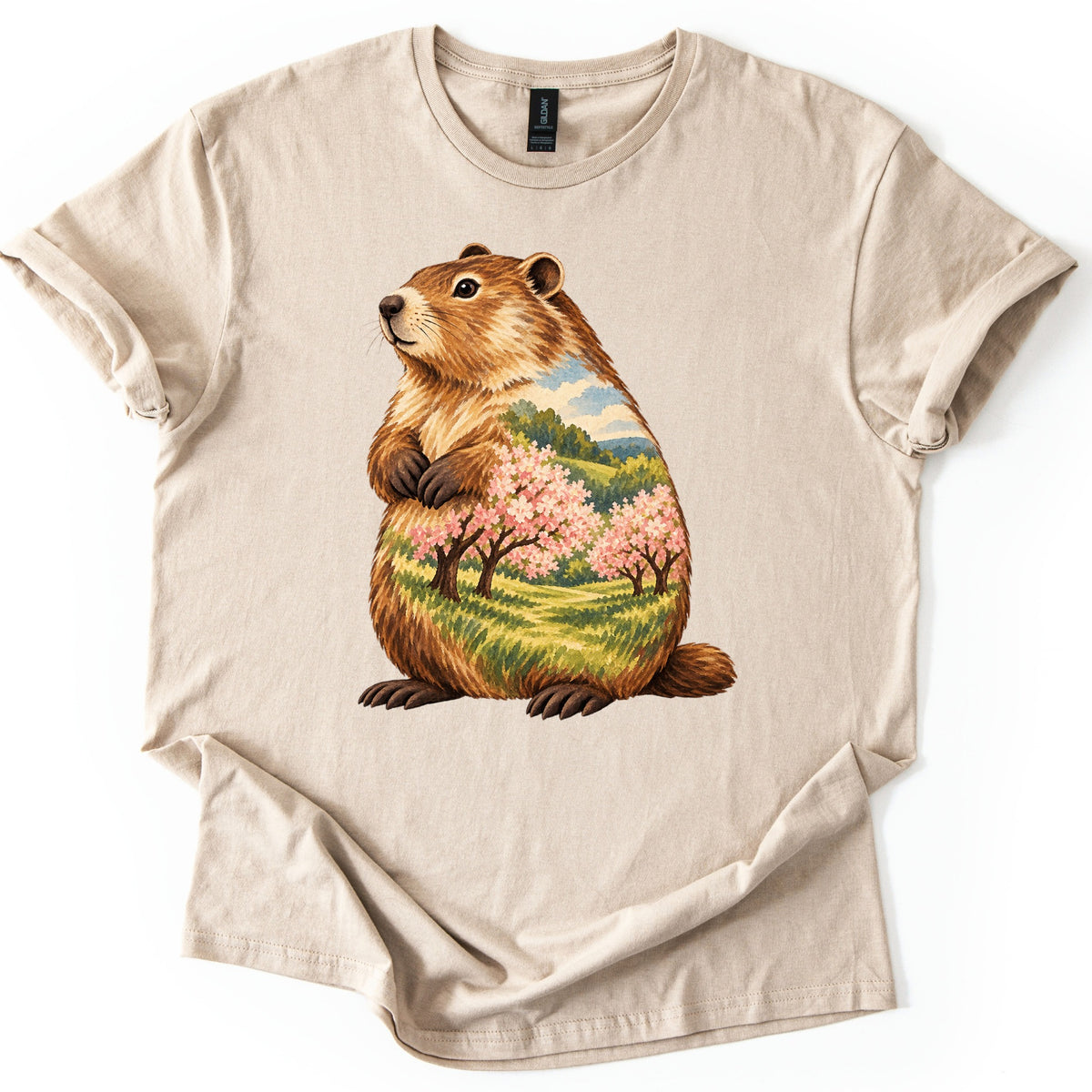 Groundhog Blossom Meadow Comfort Colors T-shirt in Sand, laid flat. A painted groundhog sits upright with paws at its chest, its body filled with a meadow of pink blossoming trees, green hills and blue sky. No words. Sleeves cuffed, on a white background.