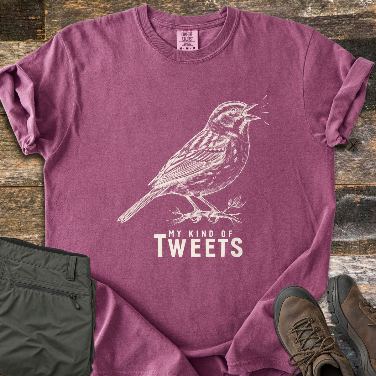 My Kind Of Tweets Comfort Colors T-shirt in Berry, laid flat. A cream sketch of a singing sparrow perched on a leafy twig, above the words My Kind Of Tweets. Shown on rustic wood with folded olive hiking pants and brown hiking boots.
