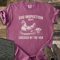 Egg Inspection Dept Comfort Colors T-shirt in Berry, laid flat. A sketched hen sits on straw beside a basket of eggs, with Egg Inspection Dept. above and Checked By The Hen below in cream. Olive cargo pants and brown hiking boots lie on a rustic wood floor.