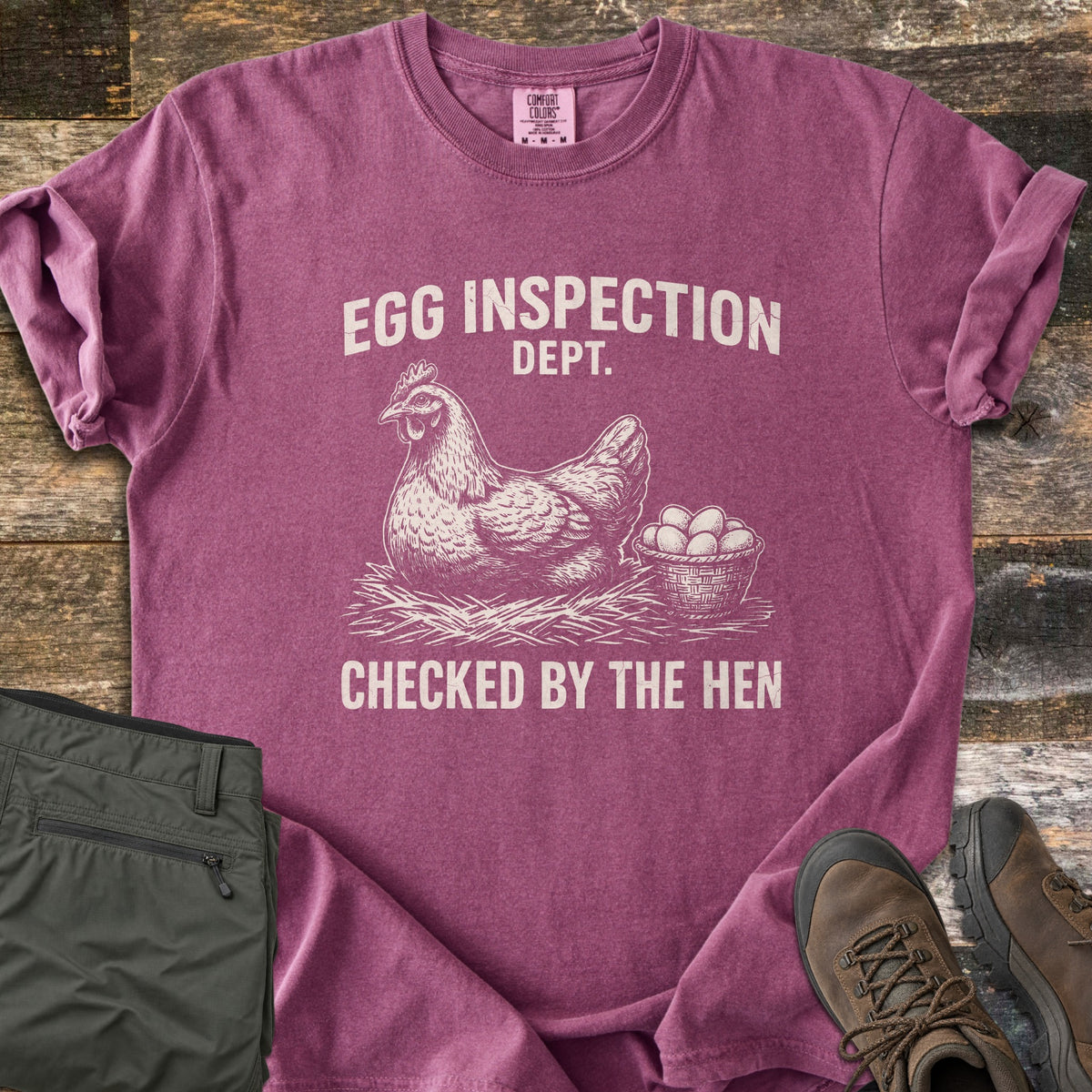 Egg Inspection Dept Comfort Colors T-shirt in Berry, laid flat. A sketched hen sits on straw beside a basket of eggs, with Egg Inspection Dept. above and Checked By The Hen below in cream. Olive cargo pants and brown hiking boots lie on a rustic wood floor.