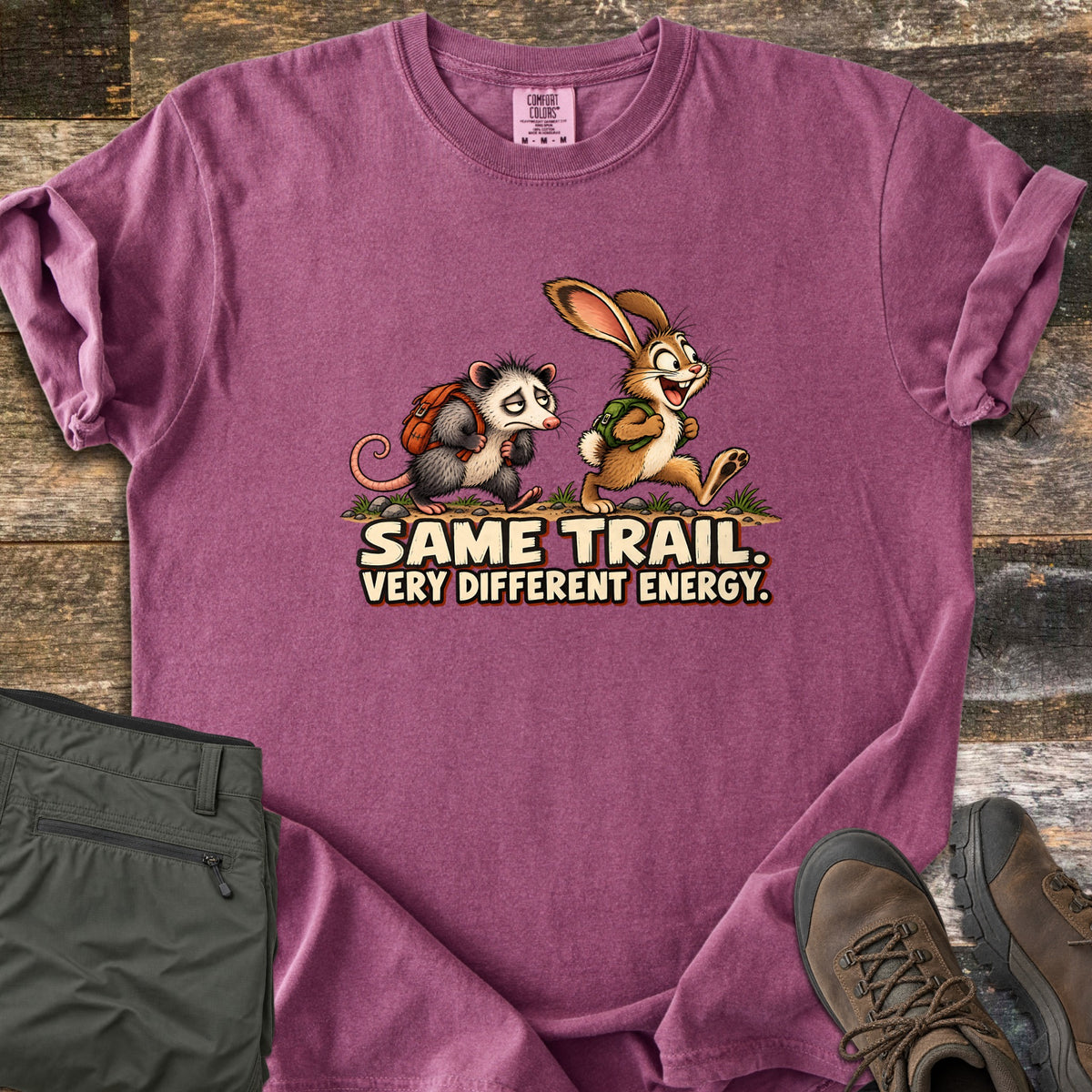 Same Trail. Very Different Energy. Comfort Colors T-shirt in Berry, laid flat. A tired cartoon opossum with an orange backpack trudges behind a grinning, bounding rabbit with a green backpack on a dirt trail, above the words Same Trail. Very Different Energy. Shown on wood with green pants and hiking boots.