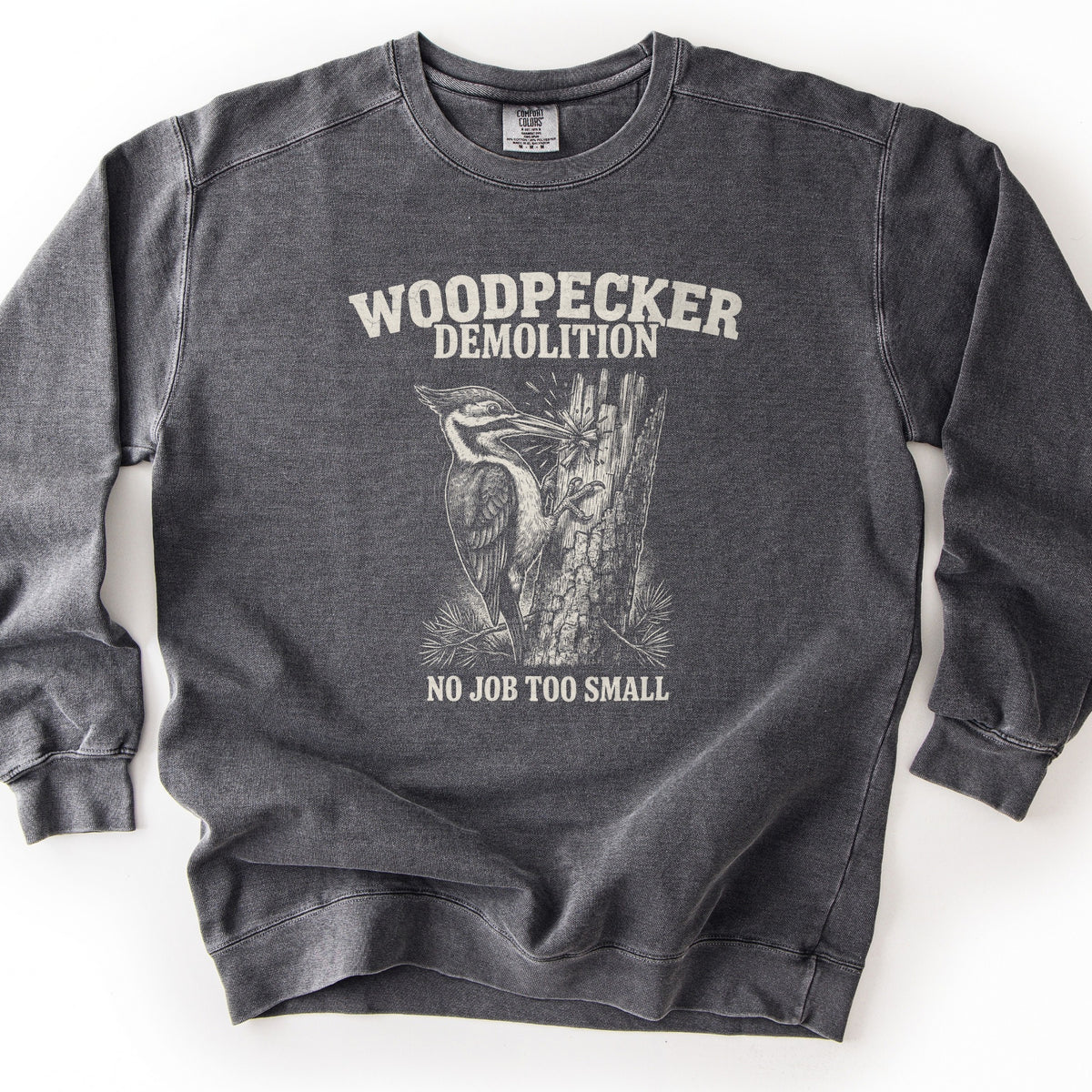 Woodpecker Demolition Comfort Colors crewneck sweatshirt in Pepper, laid flat. An etched-style drawing of a pileated woodpecker chipping splinters from a broken tree trunk, between the words Woodpecker Demolition and No Job Too Small in off-white. Shown on a plain white surface.