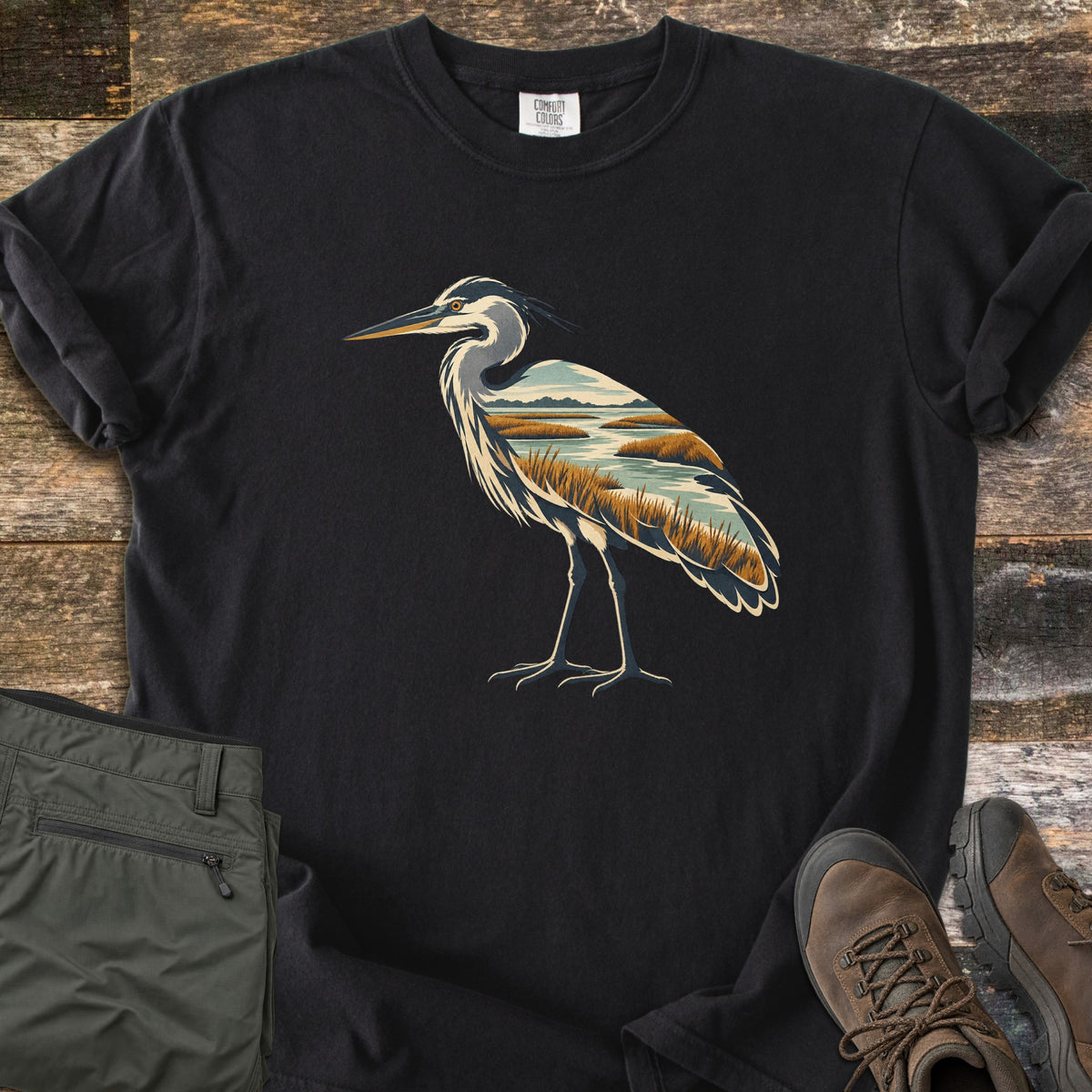 Heron Marsh Comfort Colors T-shirt in Black, laid flat. A great blue heron whose body is filled with a marsh scene of golden grasses, winding water and a cloudy sky. The shirt lies on weathered wood beside olive green pants and brown hiking boots.